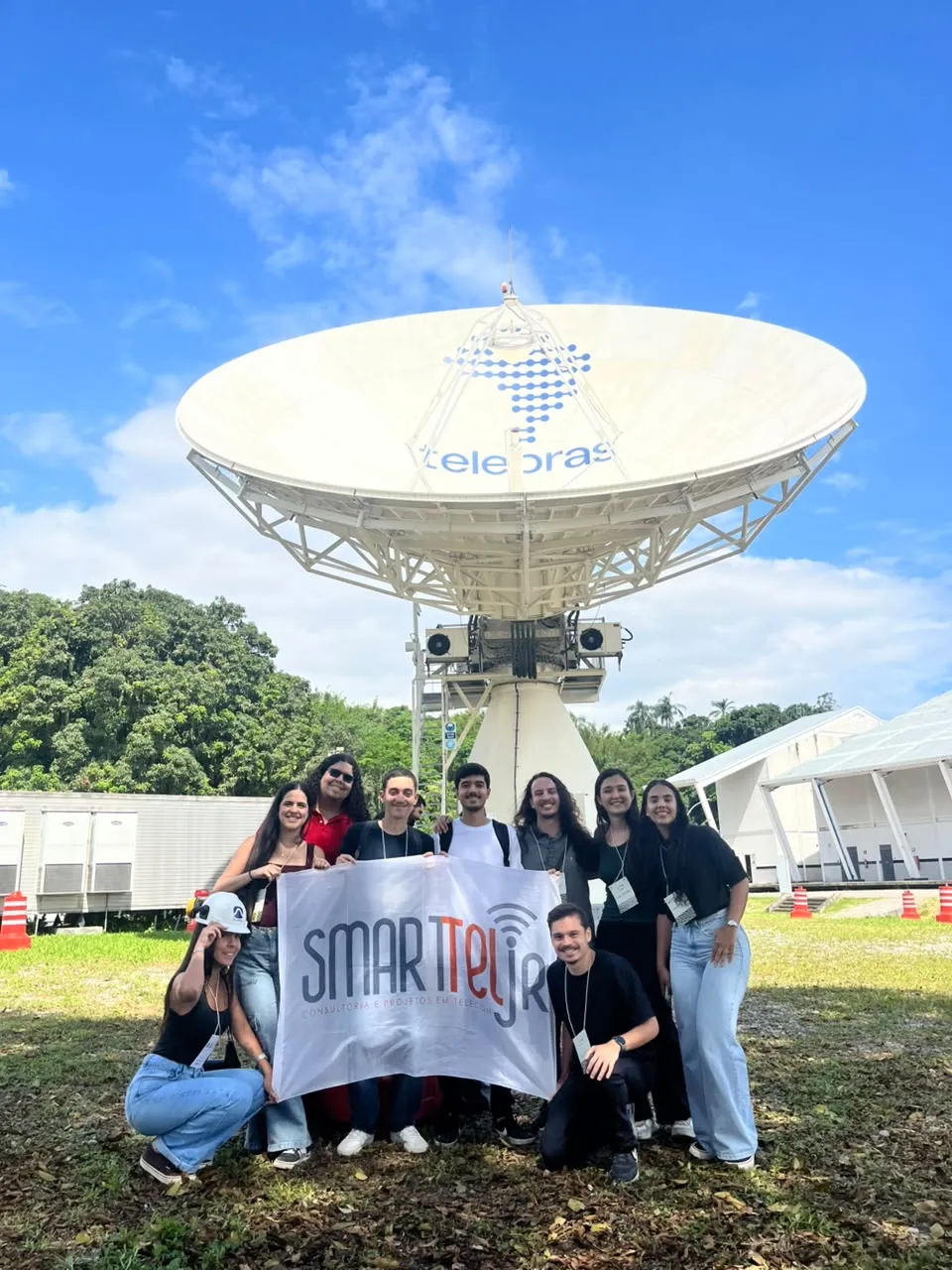Membros da SmartTel Jr com a bandeira da empresa diante de uma antena gateway do SGDC-1 no COPE-S da Telebras.